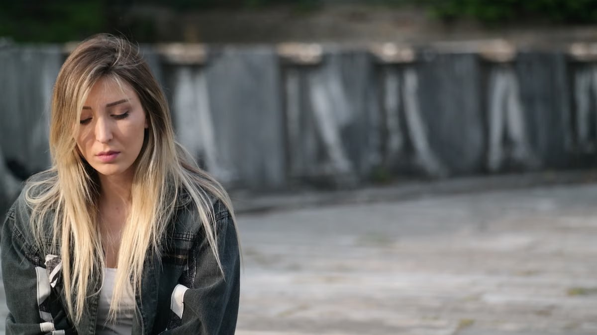 A young woman sitting outdoors, looking thoughtful and introspective.